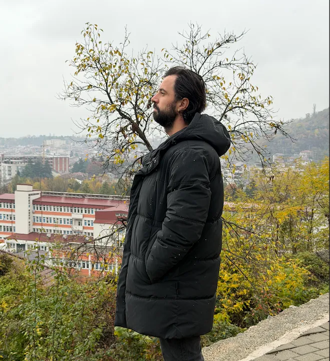 Man in a black winter coat gazing over a hillside town in autumn