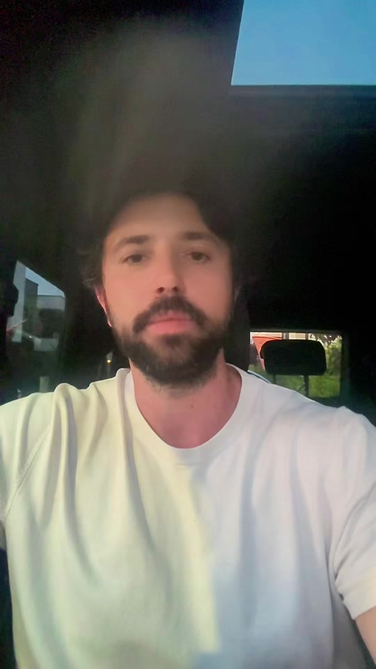 Eduard Luta in a white T-shirt speaking inside a car beneath a glass roof.
