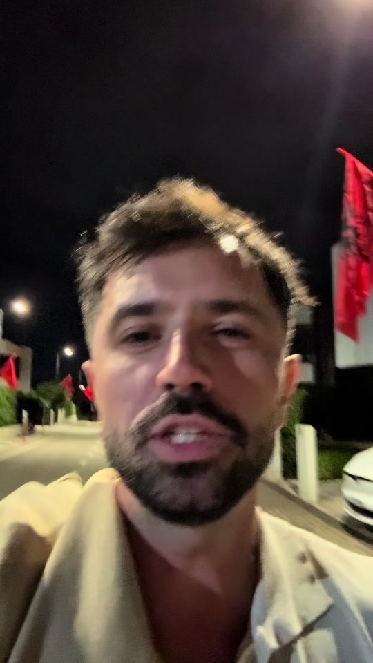 Eduard speaking while walking outdoors at night, with Albanian flags and parked cars behind him.