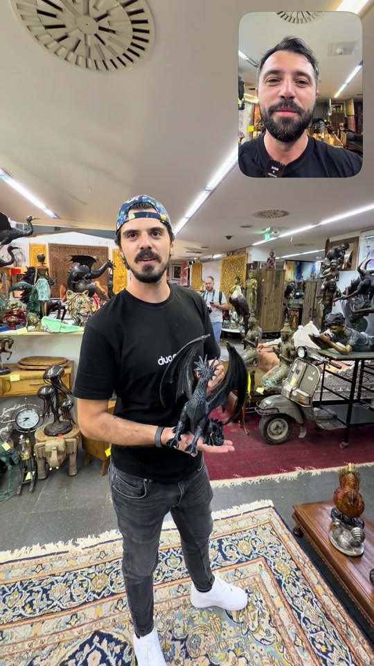 Edon holding a dark dragon figure in the Cologne shop, with rugs and decorative objects around him.