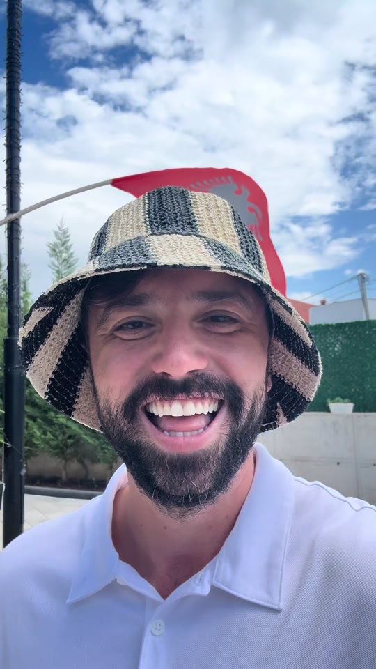 Eduard Luta smiling outdoors in a white polo and checked bucket hat, with an Albanian flag behind him.