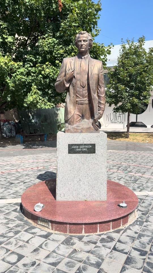 Bronze bust of Jusuf Gërvalla on a stone pedestal, surrounded by paving and trees.