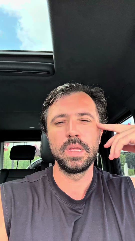 Eduard in a black sleeveless shirt inside a car, speaking with one hand near his temple.