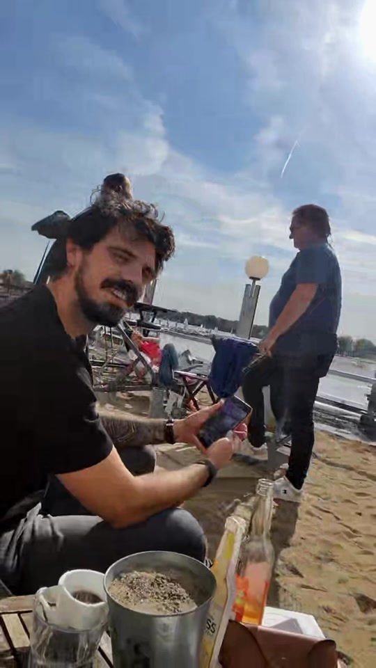 Two men beside the water at an outdoor table; one is seated holding a phone, with coffee cups in the foreground.