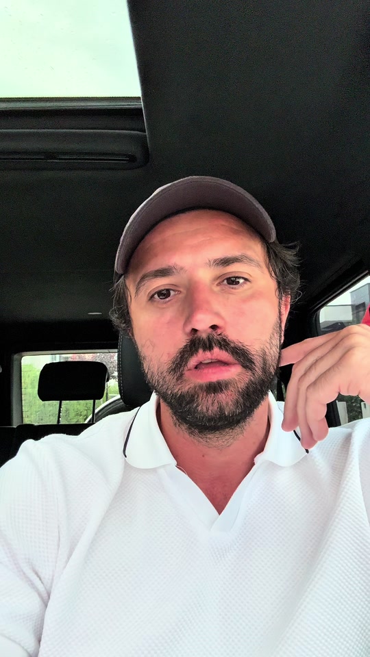 Eduard in a white polo shirt and cap inside a car, with his hand beside his face.