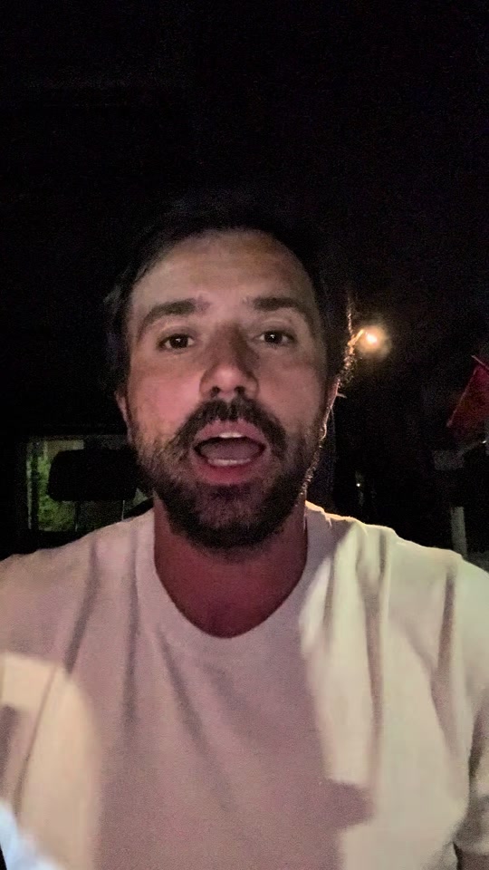 Eduard in a white T-shirt speaking to the camera at night, with a small light behind him.