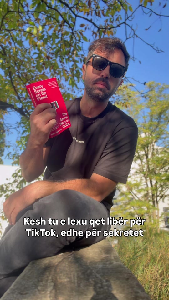 Eduard outdoors beneath a leafy tree, wearing sunglasses and holding the red Emily Baker-White book Every Screen on the Planet.