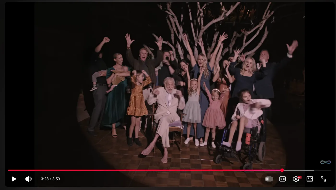 Video still of a joyful elderly man in white dancing among a cheering multigenerational family