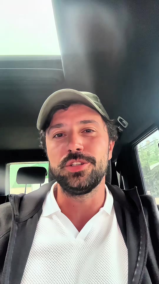 Eduard wearing a green cap, white polo and dark hoodie, recording himself inside a car.