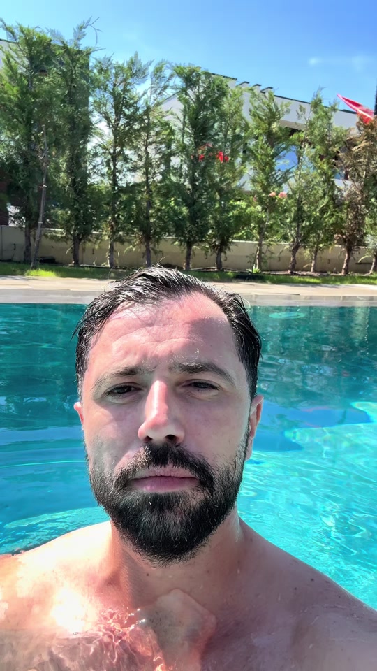 Eduard speaking from a swimming pool in sunshine, with trees behind him.